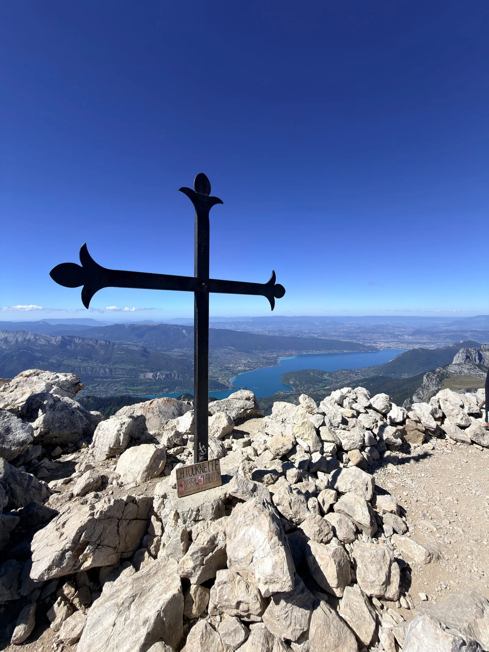 La croix du sommet de la Tournette se détache au-dessus des rochers et du lac d’Annecy.