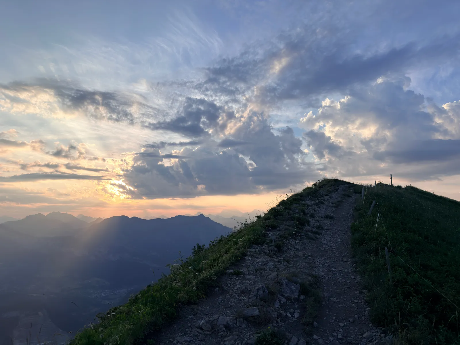 Chemin pierreux remontant une crête herbeuse sous les rayons du soleil et les nuages.