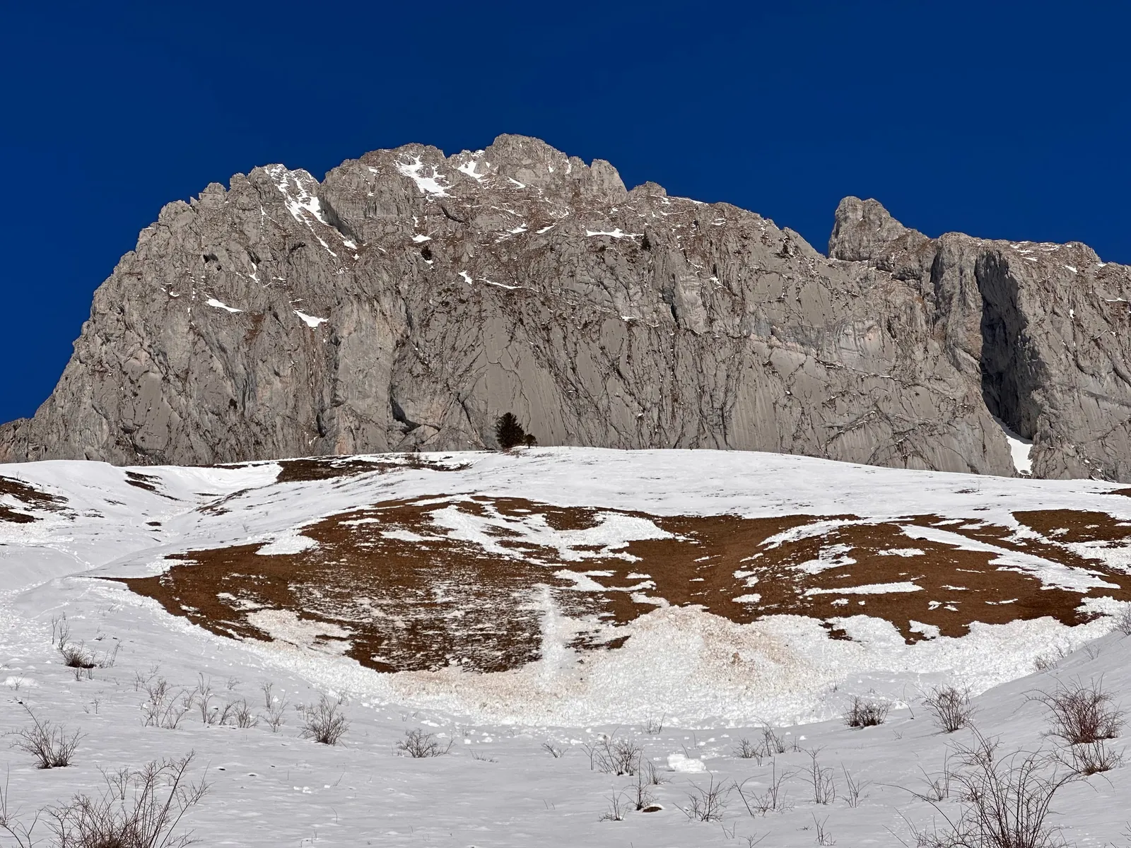 Paroi rocheuse au-dessus de pentes où la neige laisse apparaître l’herbe.